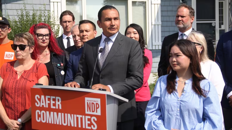 Photo of Wab Kinew speaking at press conference in August 2023. The sign in front of him says "safer communities."