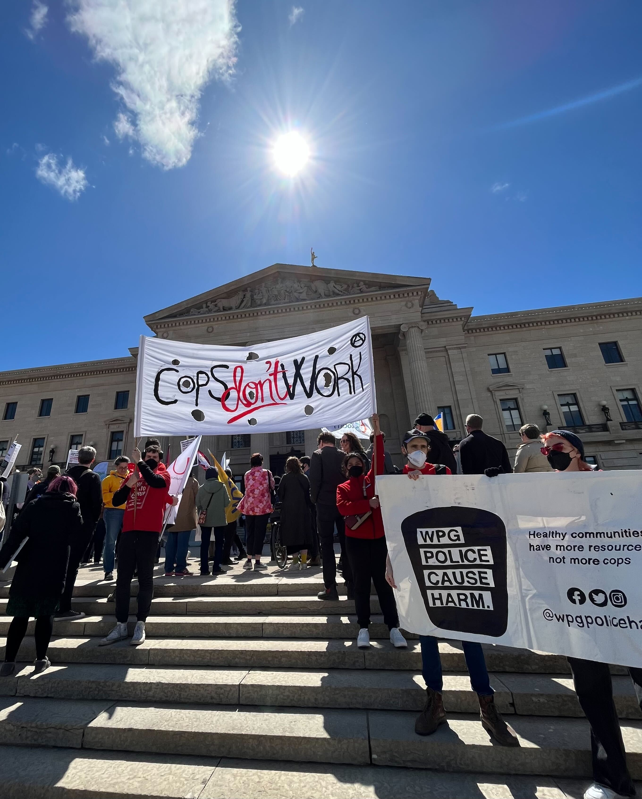 A photo taken on the lower steps of the Manitoba Legislature beneath a bright blue sky and sun. In the middle is a large banner that reads "Cops Don't Work." On the right is a WPCH banner.