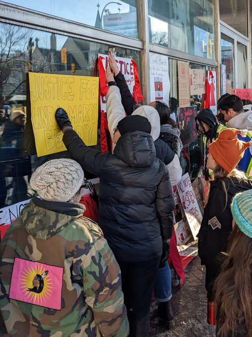 People tape signs to the windows of police HQ. The closest sign is yellow and in black font reads “justice for Linda Mary Beardy.”