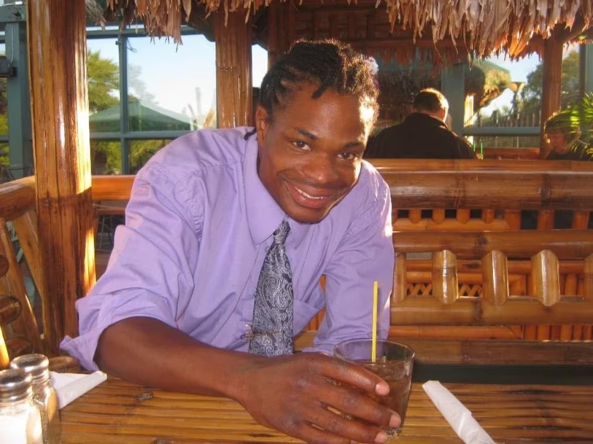 Photo of Dustin sitting at a restaurant, holding a drink, and smiling. He is wearing a lavender coloured shirt with a tie.