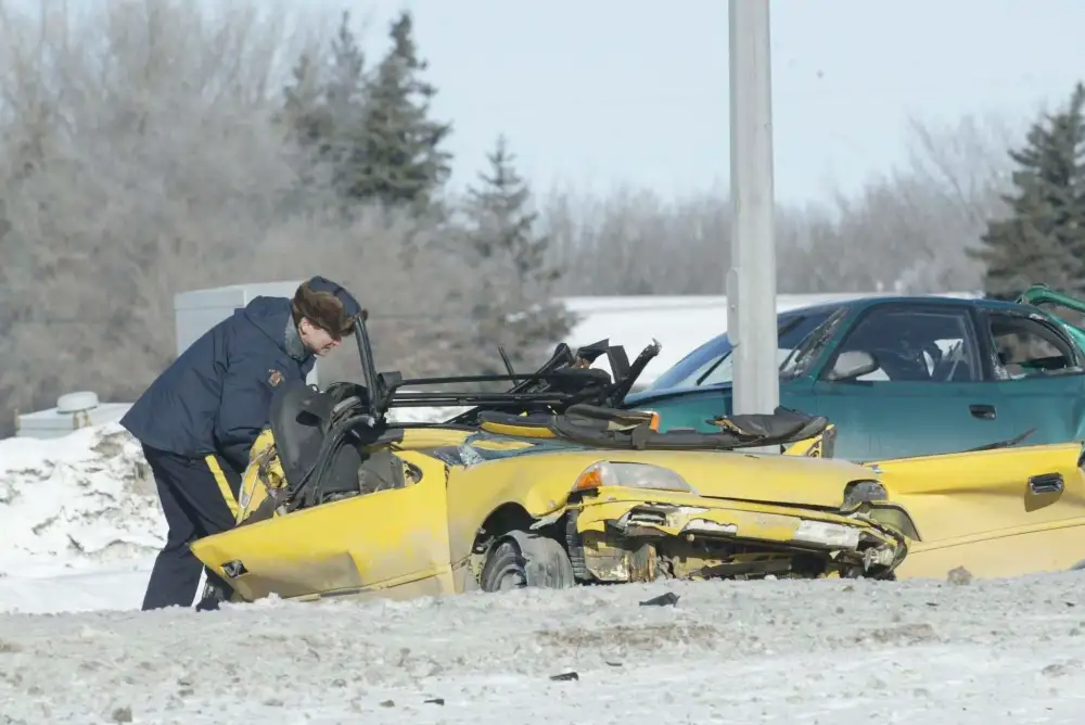 A photo of a crumpled up yellow car and a police officer peering into it. A green truck or SUV is behind it.